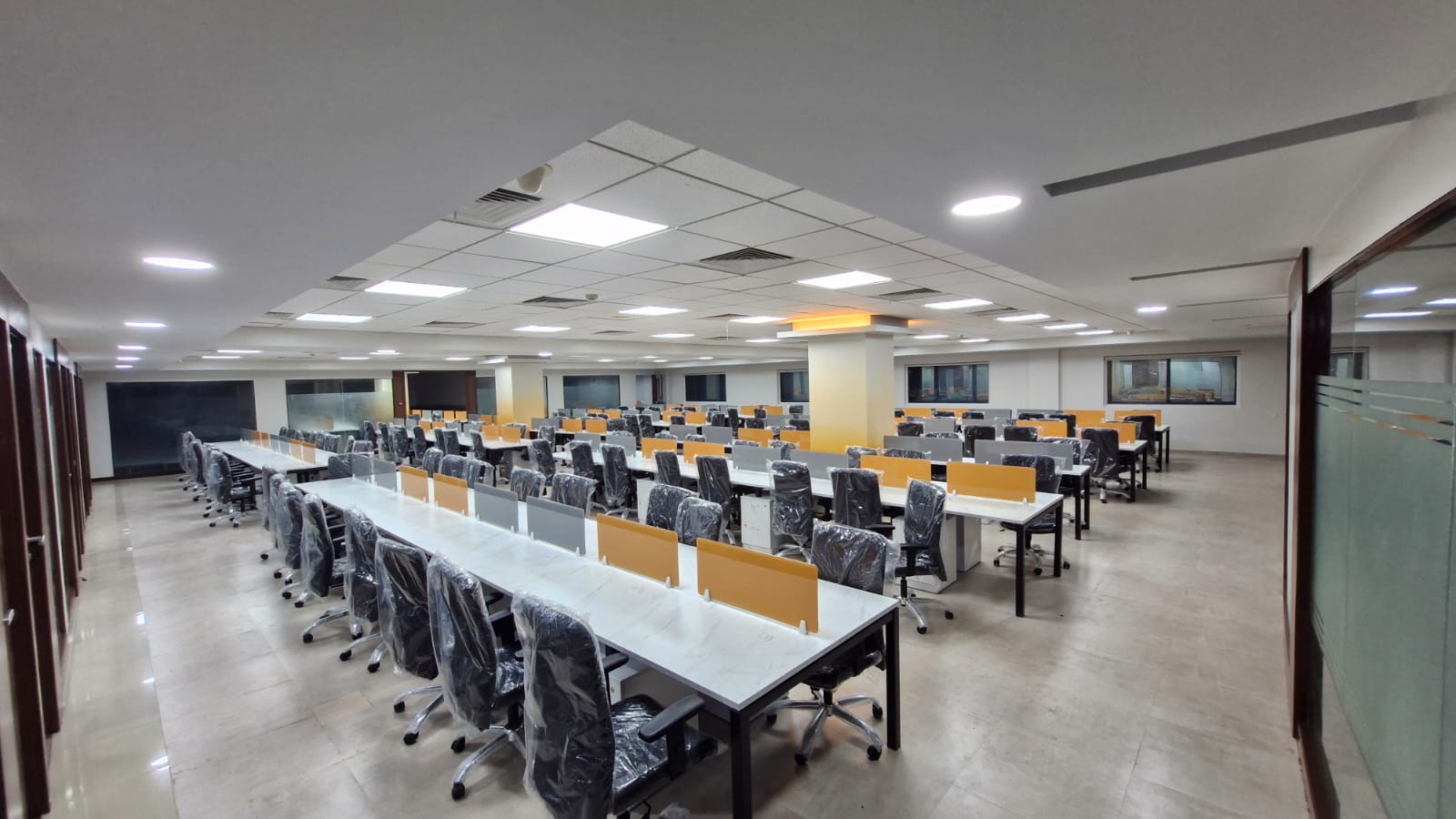 Open-plan office with long rows of white workstations, orange screens and glass-walled cabins along one side