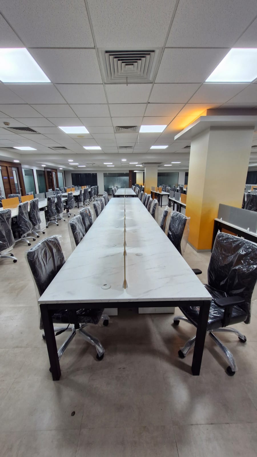 A long double row of white workstations with black mesh chairs under a tiled ceiling