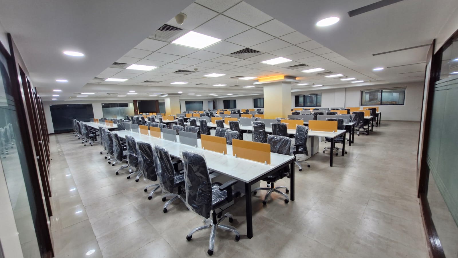Rows of white workstations with orange desk screens beside glass-walled cabins