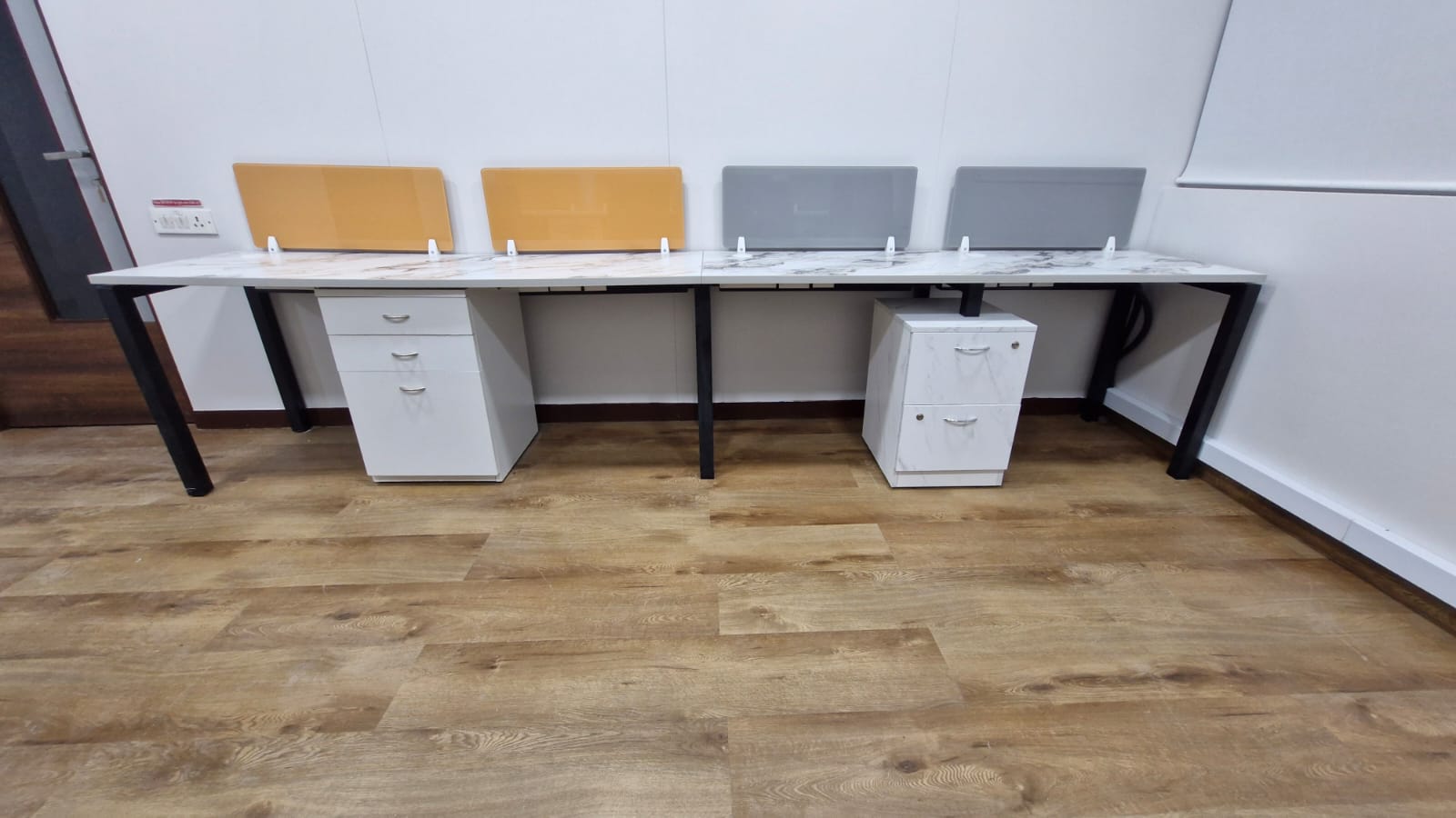 Compact white workstations with orange and grey desk screens and white pedestals on a wood-look floor