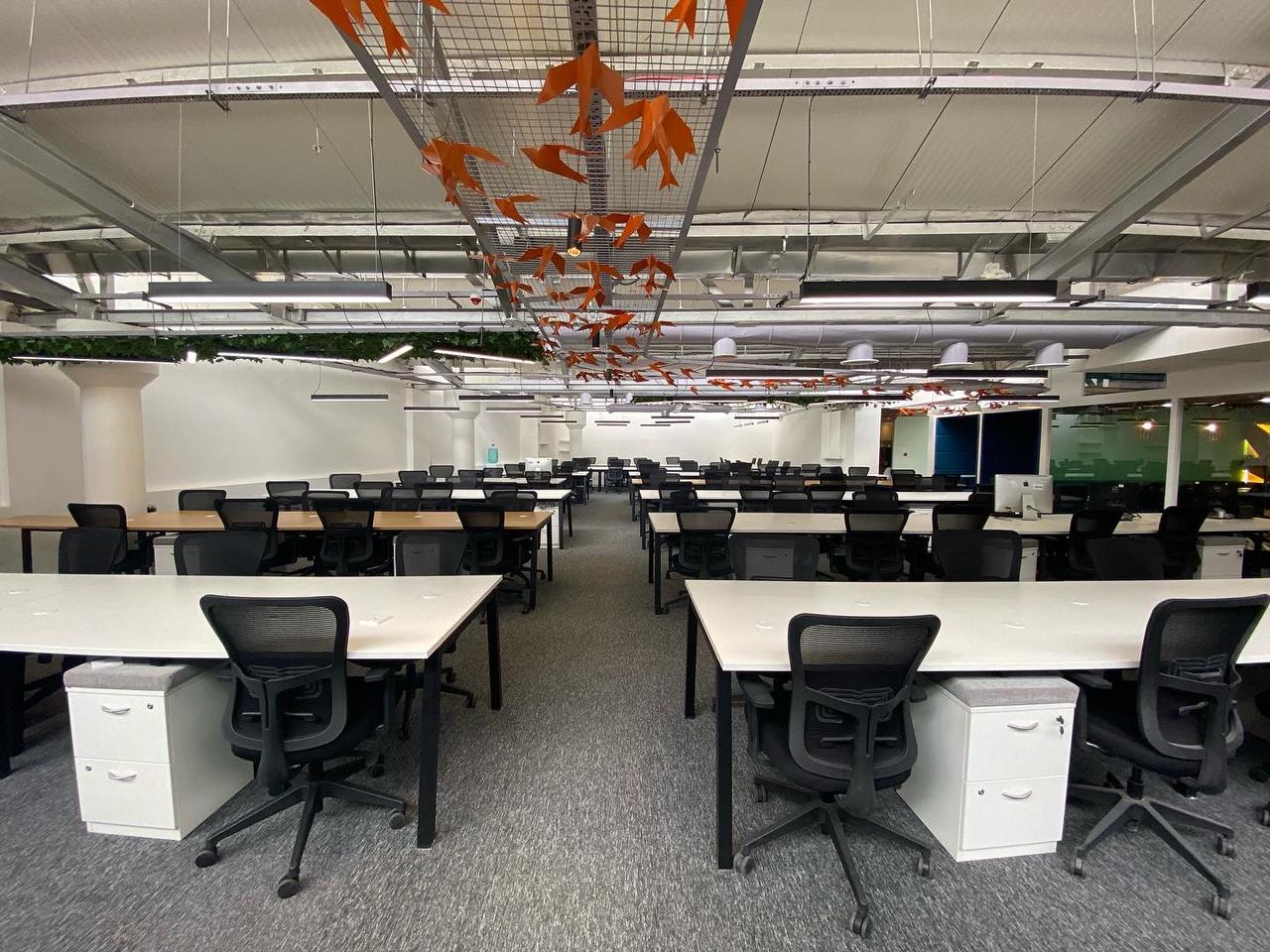 Open-plan office with rows of white-top workstations beneath a ceiling installation of orange paper birds